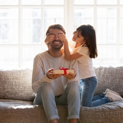 Small daughter congratulate daddy with father day, people sitting on sofa kid covering dad eyes with hands prepare for him gift box with surprise. Life events celebrating, love and attention concept